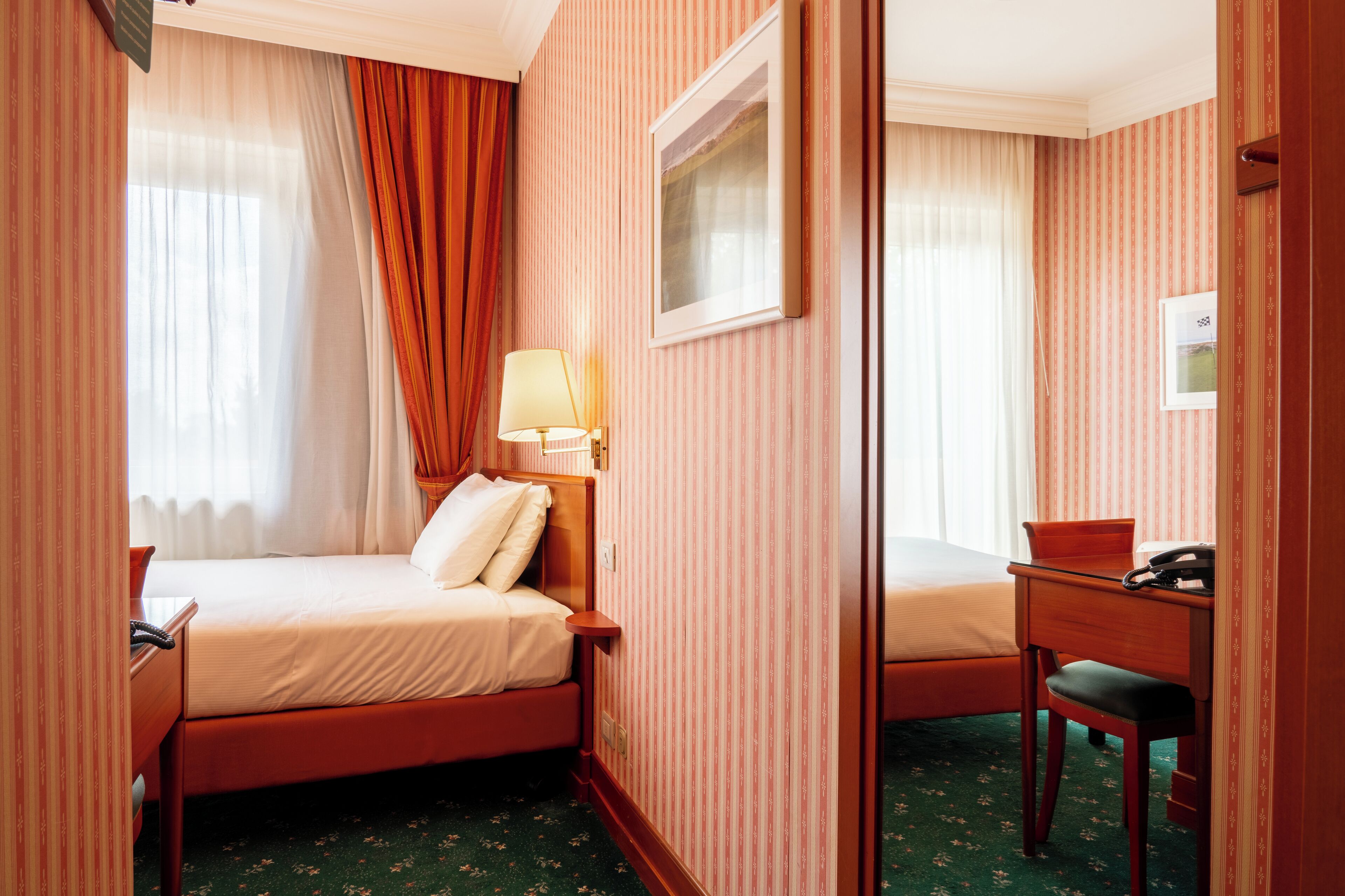 A hotel room with a bed, a desk, a chair, a lamp, and a mirror.