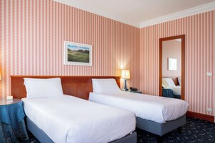 Habitación de hotel con dos camas individuales, cabecera de madera, mesita de noche con lámpara, un cuadro enmarcado en la pared y un espejo.