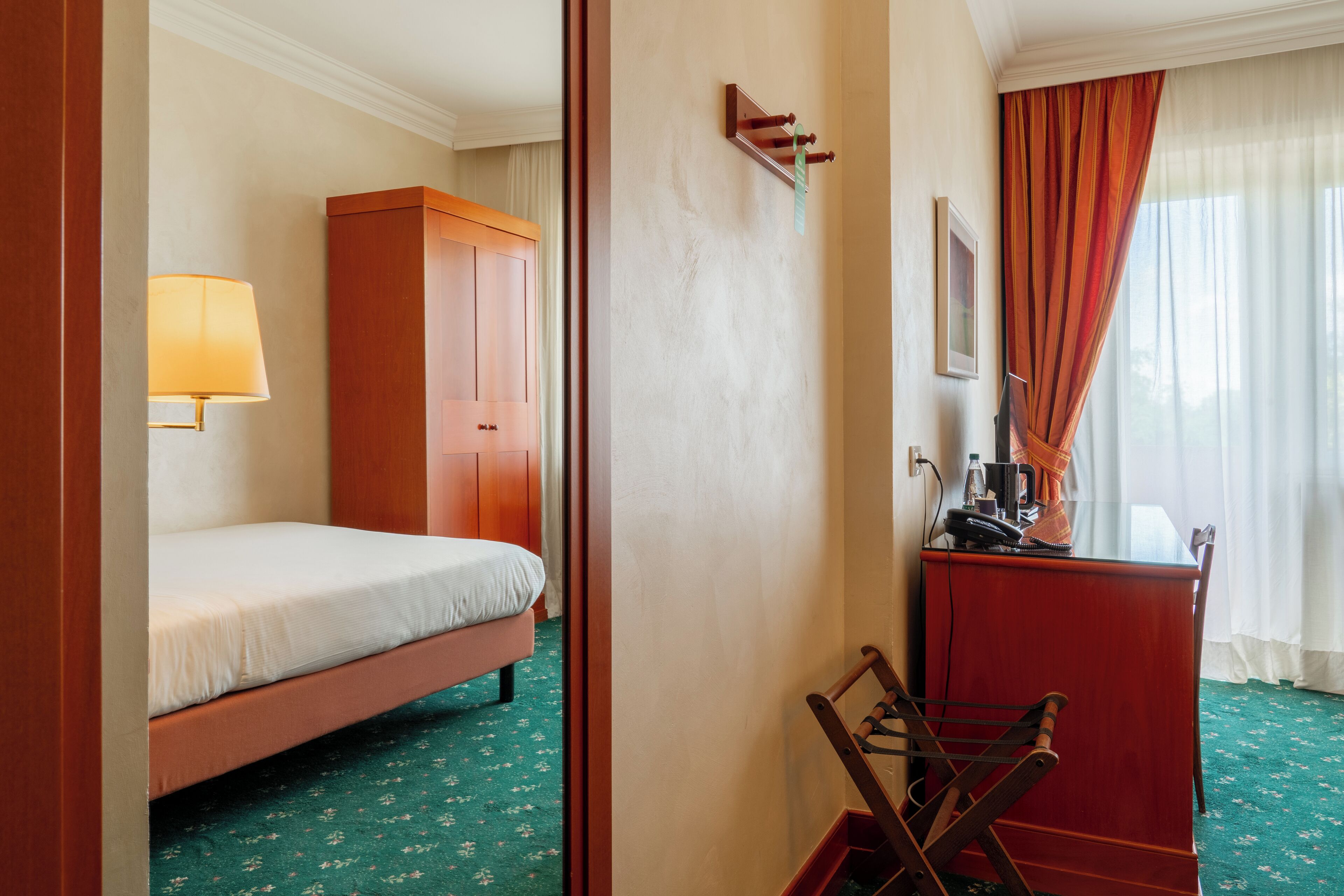 A hotel room with a bed, a desk, a chair, a lamp, and a wardrobe.