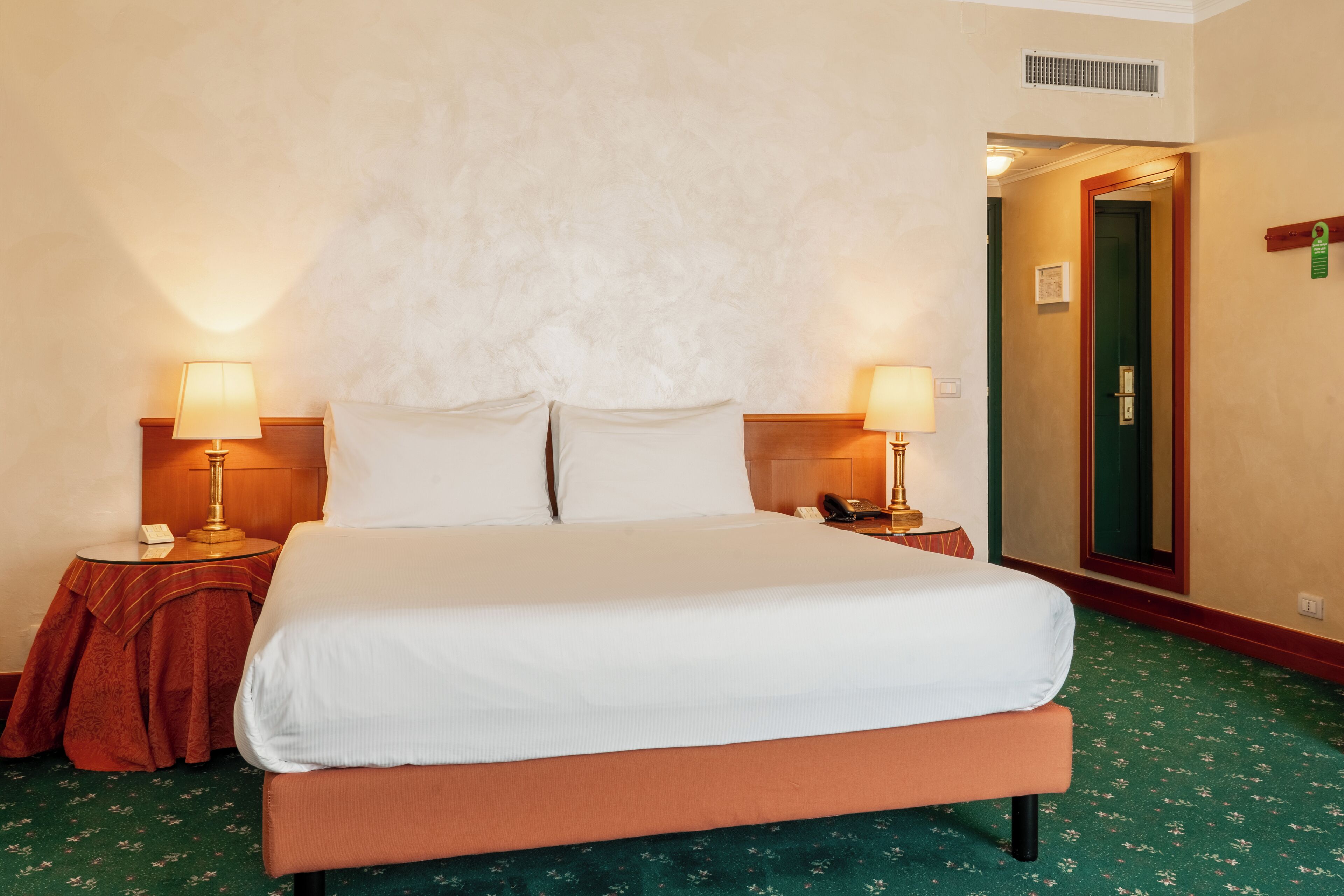 Ein Hotelzimmer mit einem großen Bett, zwei Nachttischen mit Lampen, einem grünen Teppich und einer Tür zu einem weiteren Zimmer.