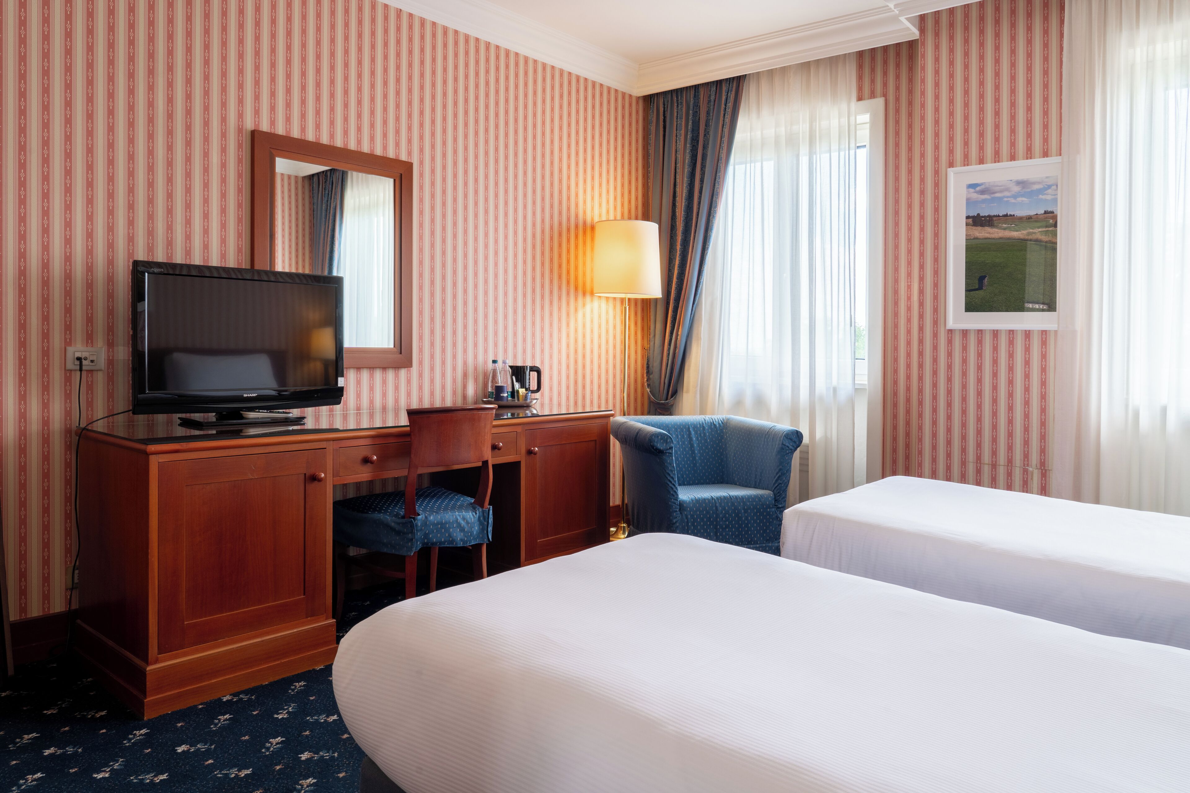Une chambre d’hôtel avec un papier peint à rayures, une télévision, un bureau avec une lampe, deux lits et une chaise.