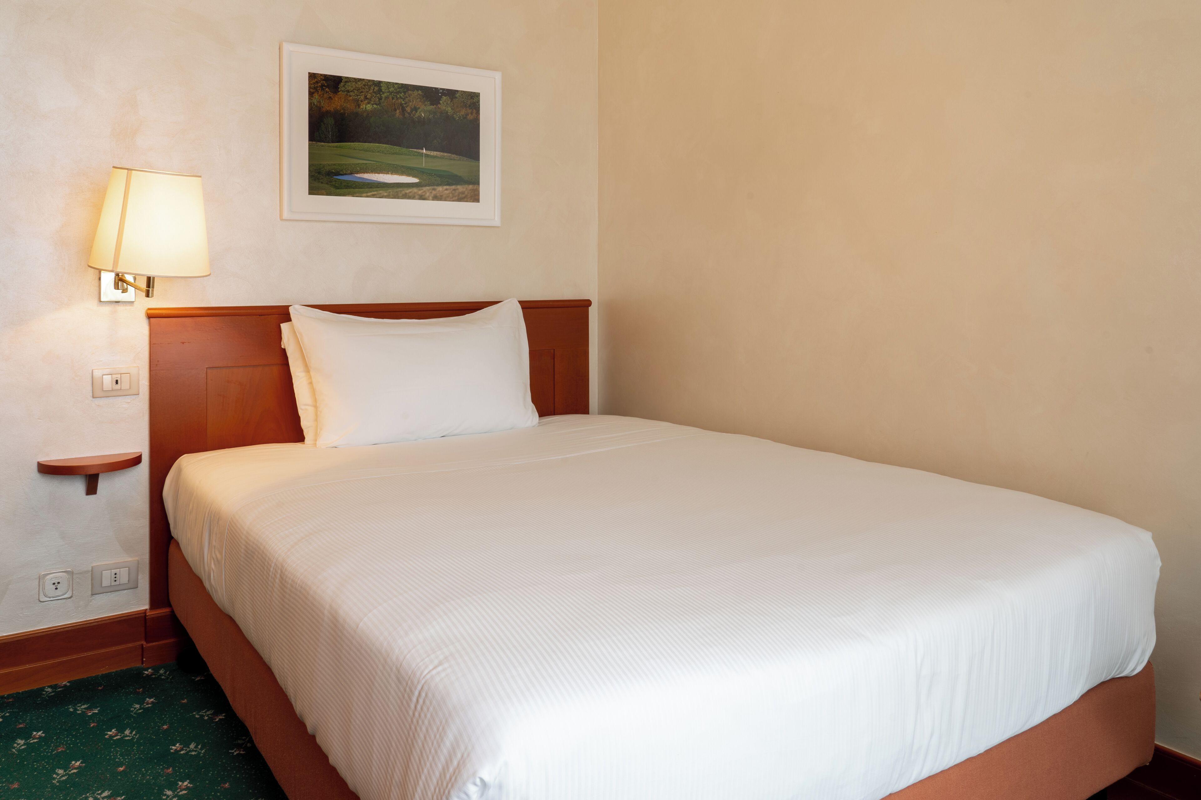 Un lit bien fait, avec une couette blanche et des oreillers, une tête de lit en bois, une lampe fixée au mur et un tableau représentant un terrain de golf.