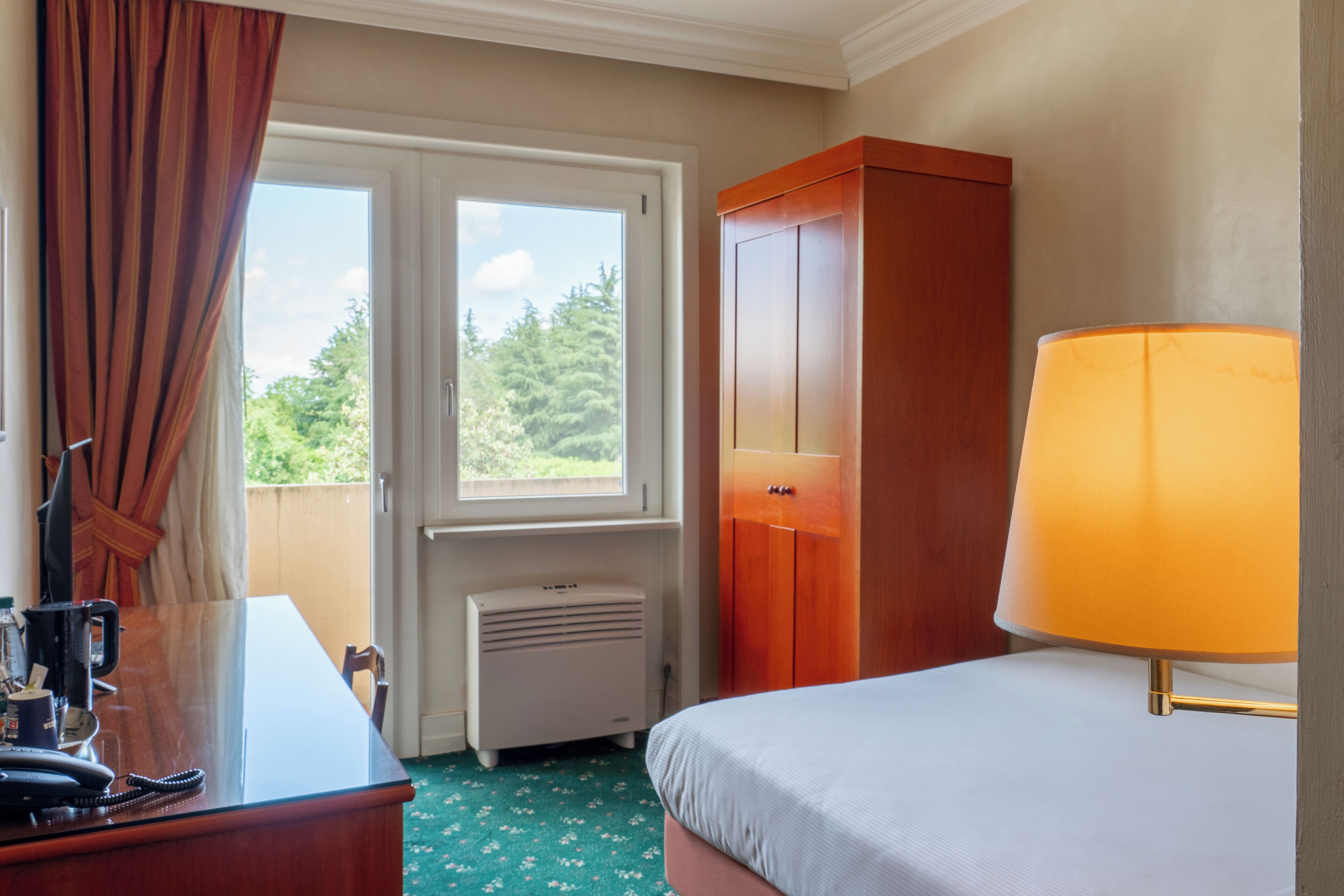Une chambre d’hôtel comprenant un lit, un bureau avec un téléphone, une lampe, une armoire et une fenêtre donnant sur des arbres.