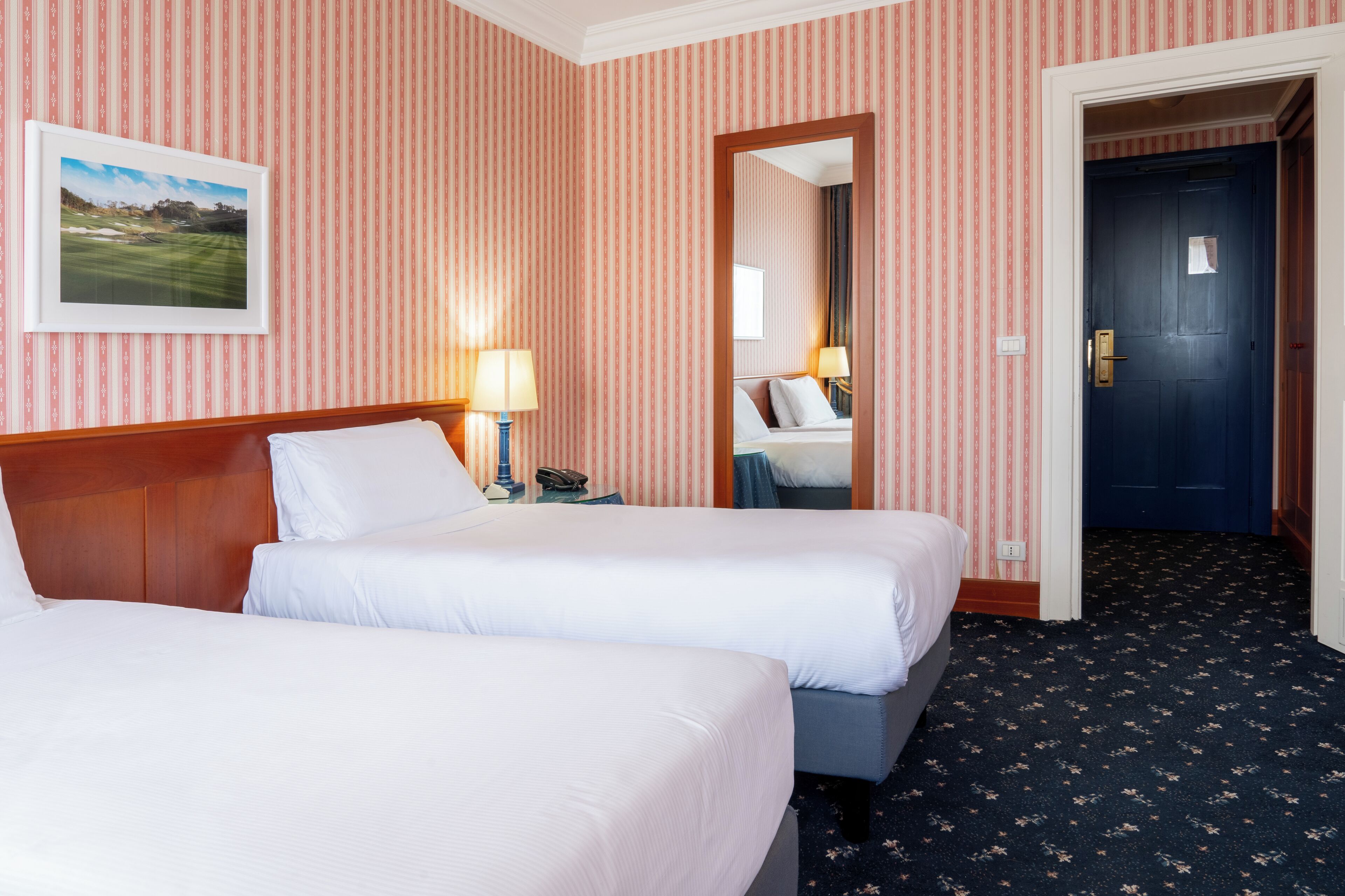 Ein Hotelzimmer mit zwei Betten, gestreifter Tapete, einem gerahmten Bild an der Wand, einem Spiegel und einem dunkel teppichbedeckten Boden.