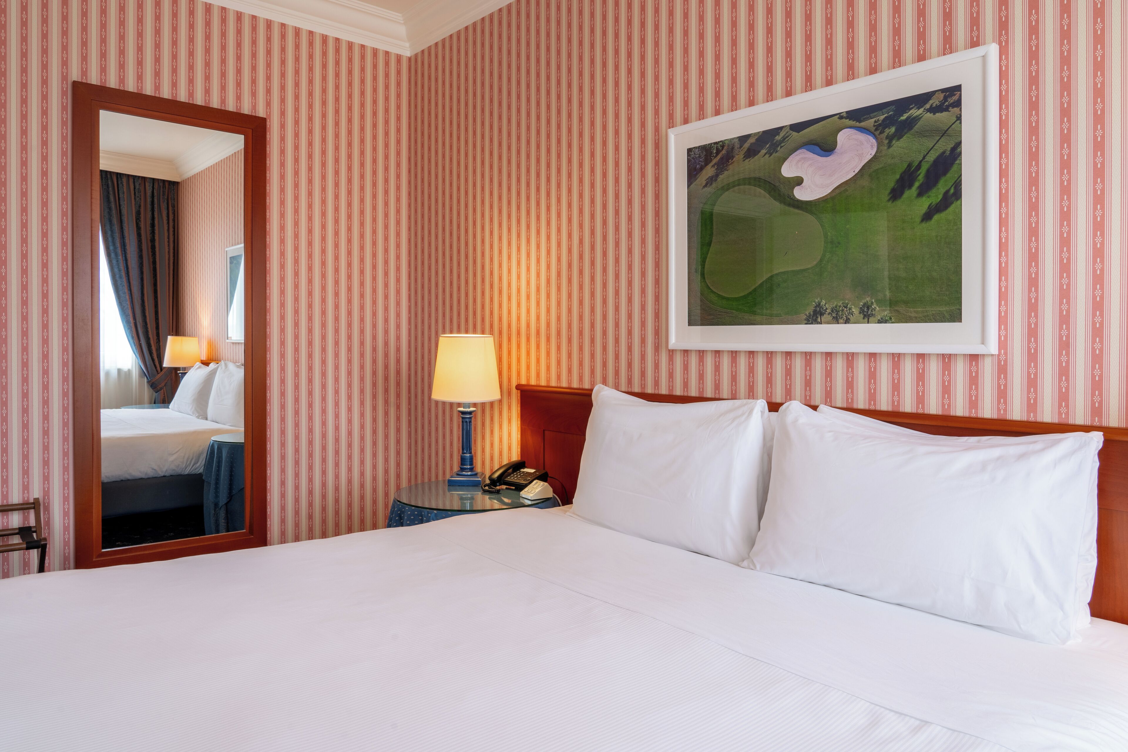 Une chambre d’hôtel avec un grand lit, une table de chevet avec une lampe, un tableau encadré au mur et un miroir qui reflète la pièce.