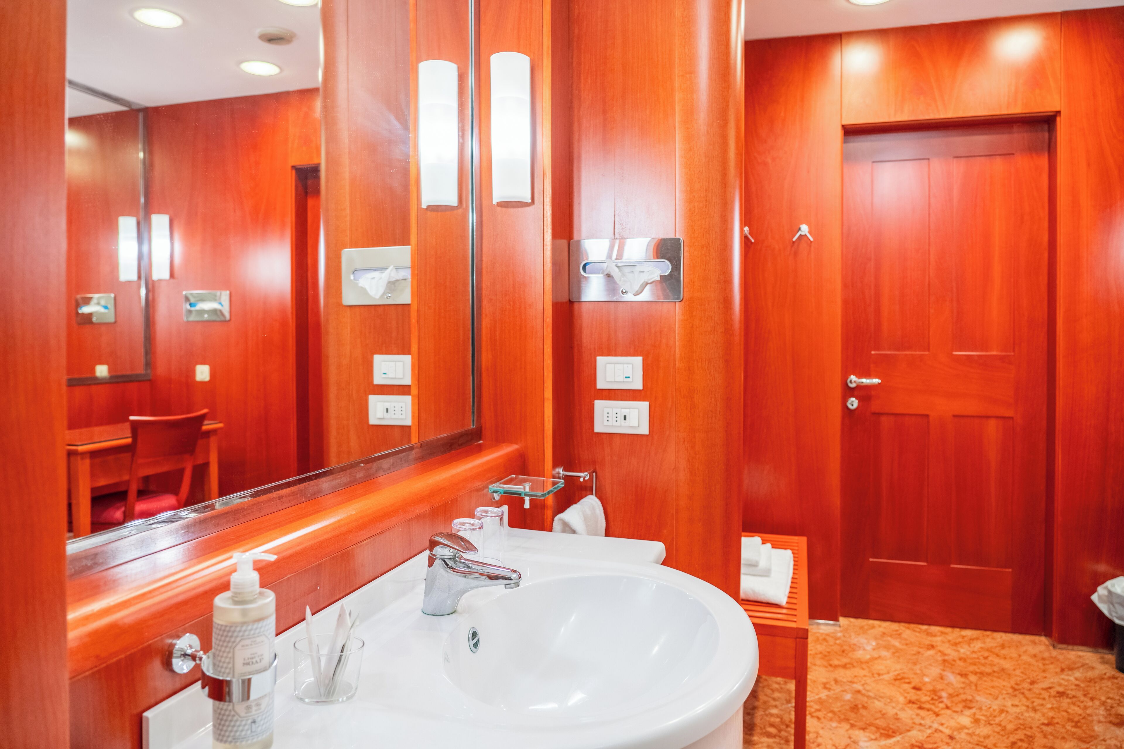 Un baño de hotel con un lavamanos grande de color blanco, paredes de madera, un espejo y una puerta.