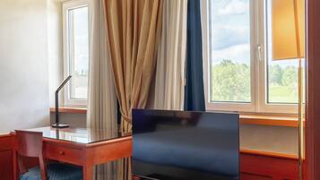 A hotel room with a desk, chair, TV, and large windows with curtains.