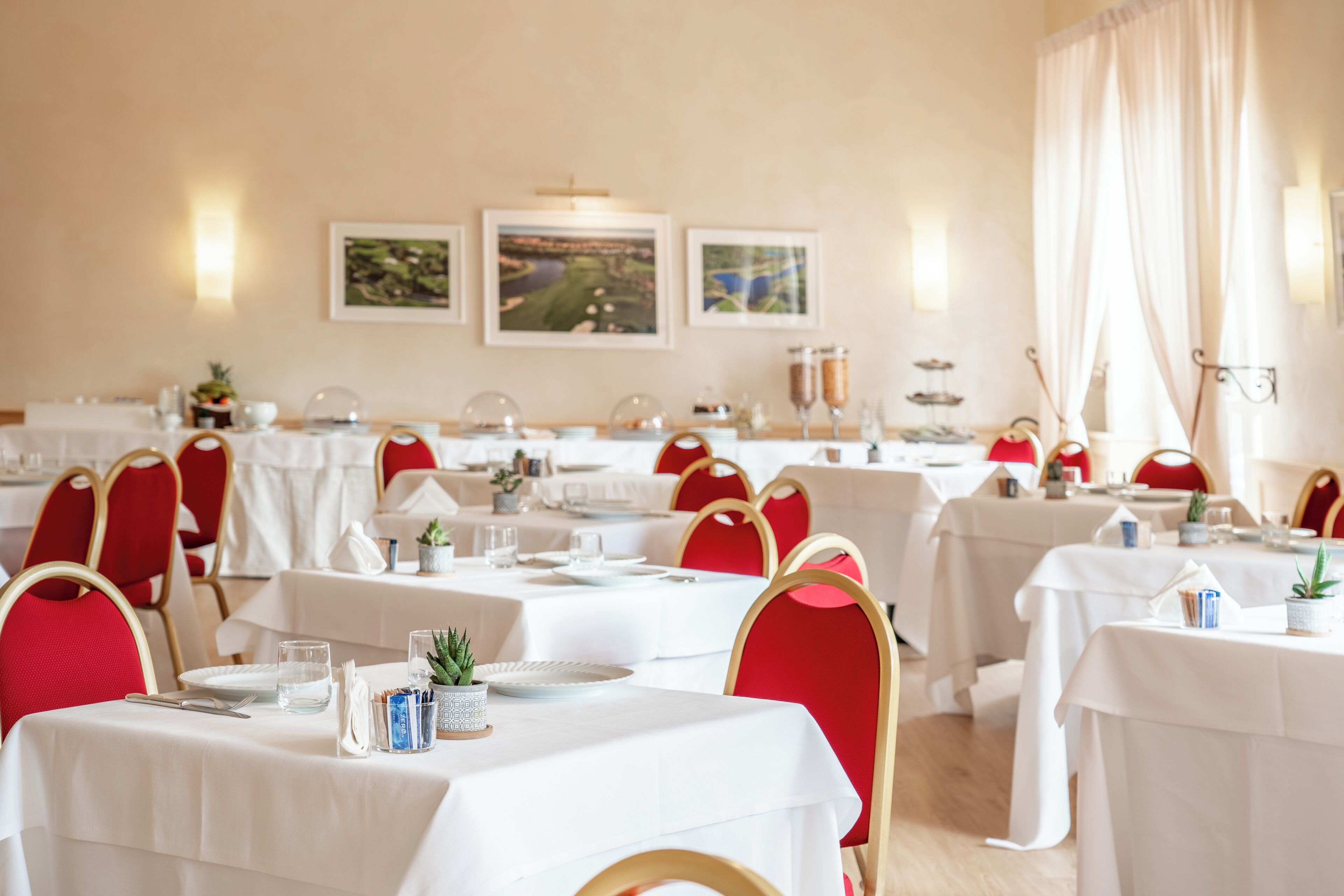A restaurant with multiple tables set for dining, red chairs, and framed pictures on the wall.