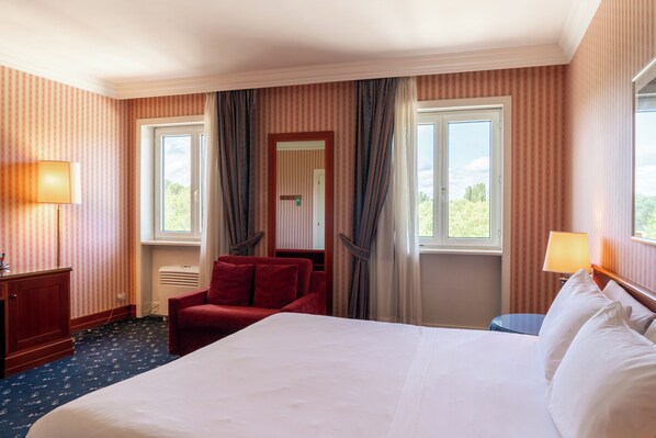 A hotel room with a large bed, a red armchair, a desk with a lamp, and a view of greenery outside. - Golf Hotel Cavaglià (Cavaglia)