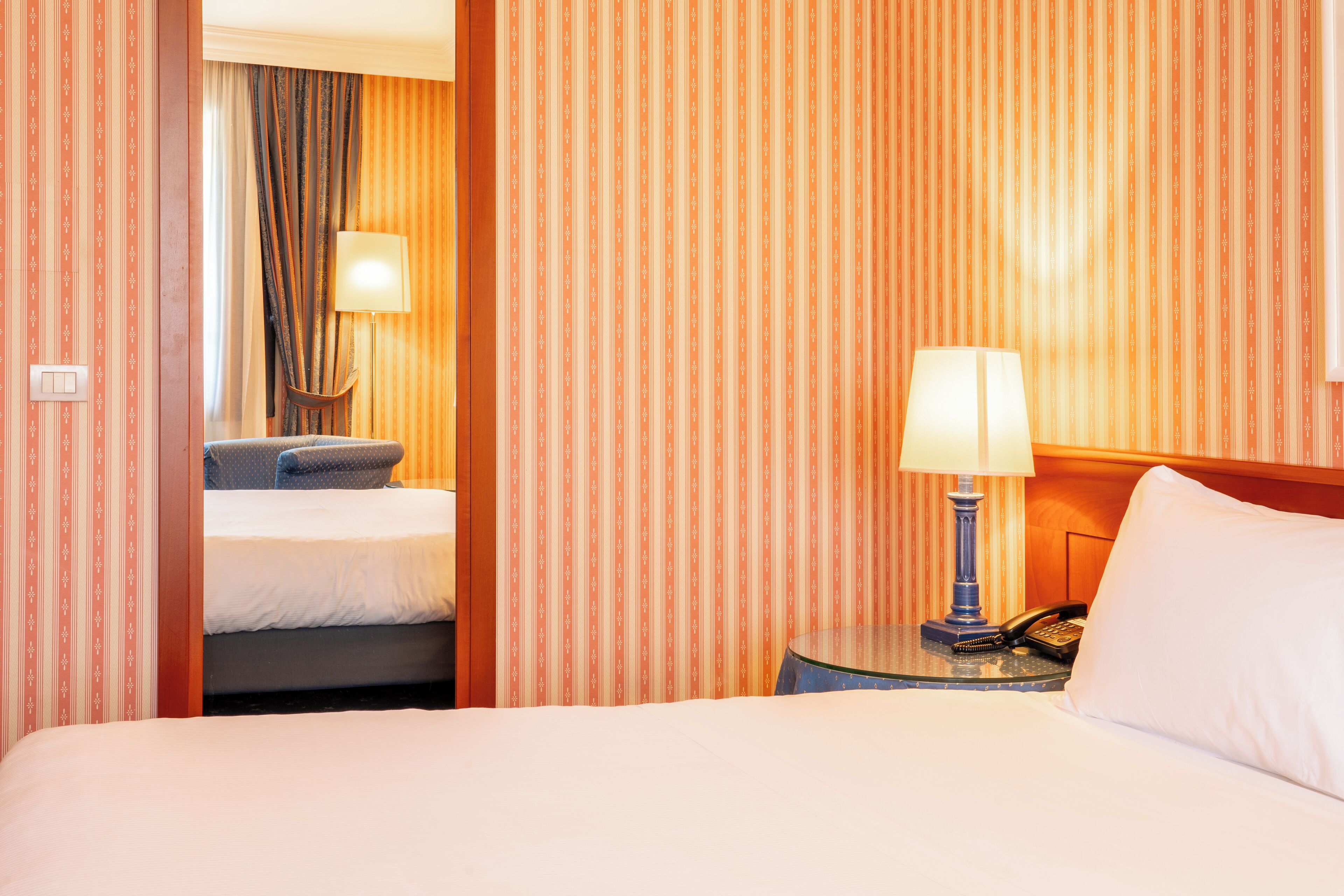 Habitación de hotel con papel tapiz a rayas, una cama, una mesita de noche con lámpara y un teléfono.