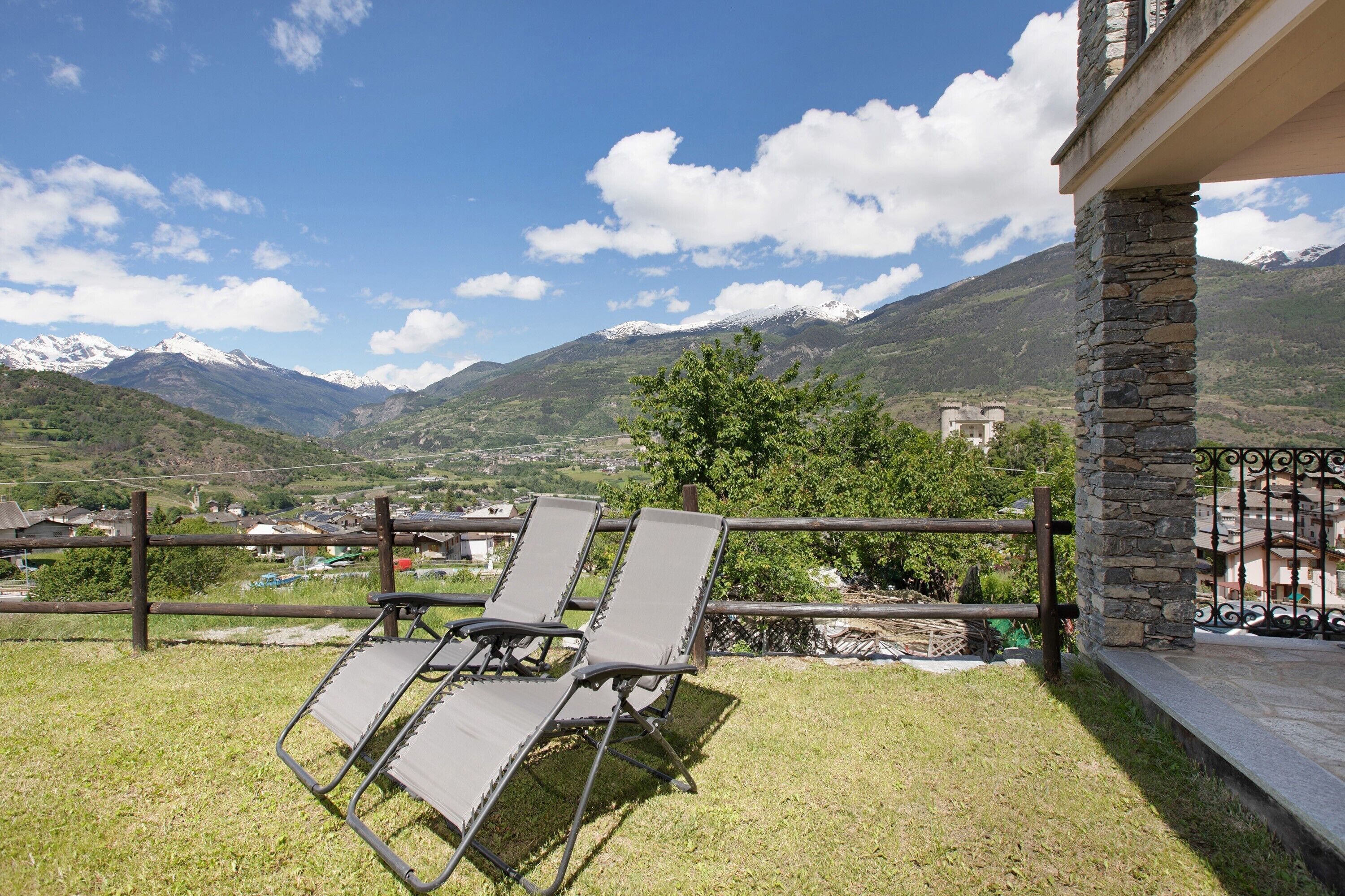 A balcony with two lounge chairs, a stone pillar, and a view of mountains and a village.