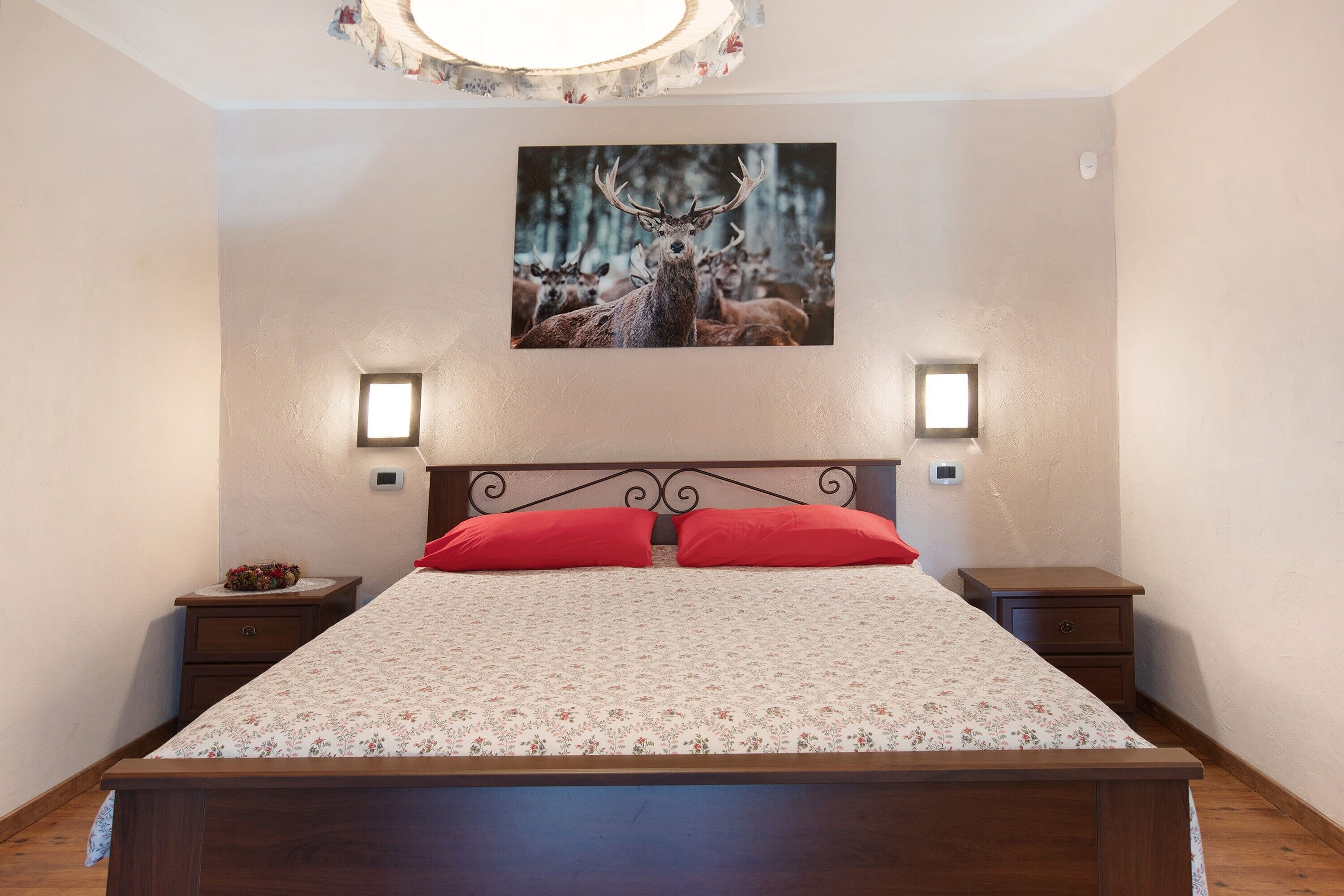 A bedroom with a wooden bed, red pillows, two bedside tables, a nightstand, a wall-mounted picture of deer, and two wall-mounted lights.