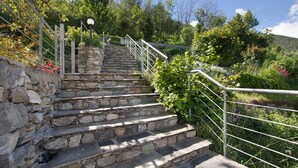 Un escalier en pierre, agrémenté de rampes métalliques, mène au sommet d’une colline, le tout étant entouré de verdure et d’arbres.