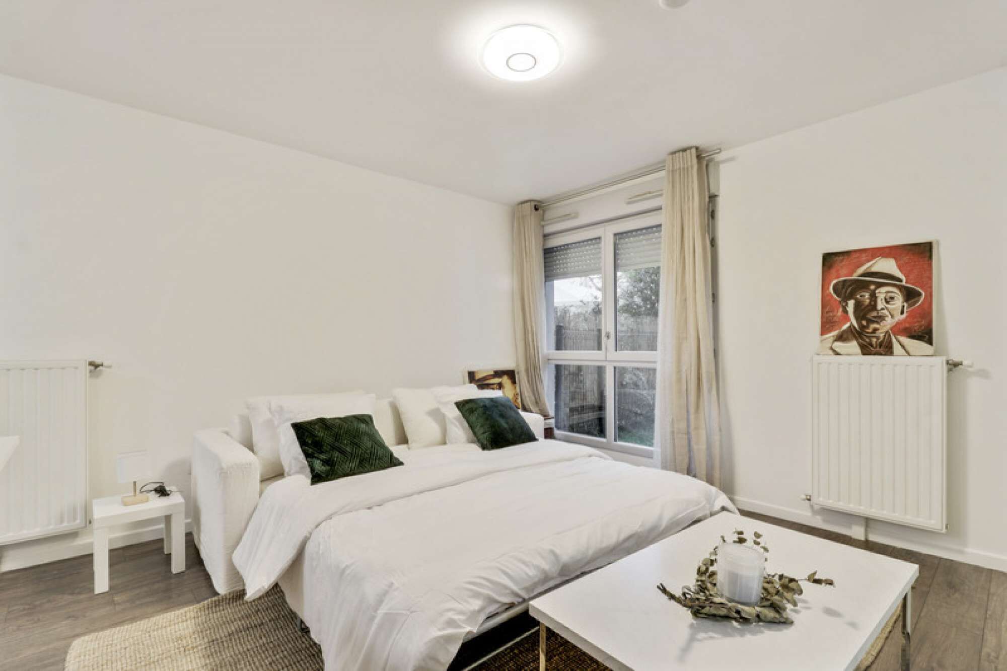 A bedroom with a bed, a bedside table, a window with curtains, a radiator, and a painting on the wall.