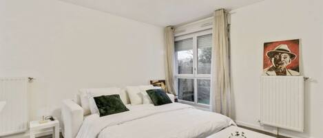 A bedroom with a bed, a bedside table, a window with curtains, a radiator, and a painting on the wall.