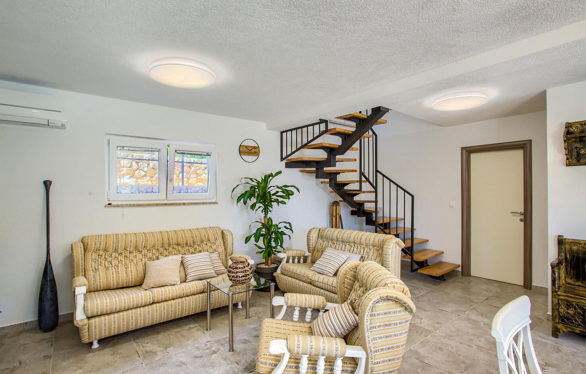 A living room with a striped sofa, armchairs, a coffee table, a potted plant, a staircase, and a door.