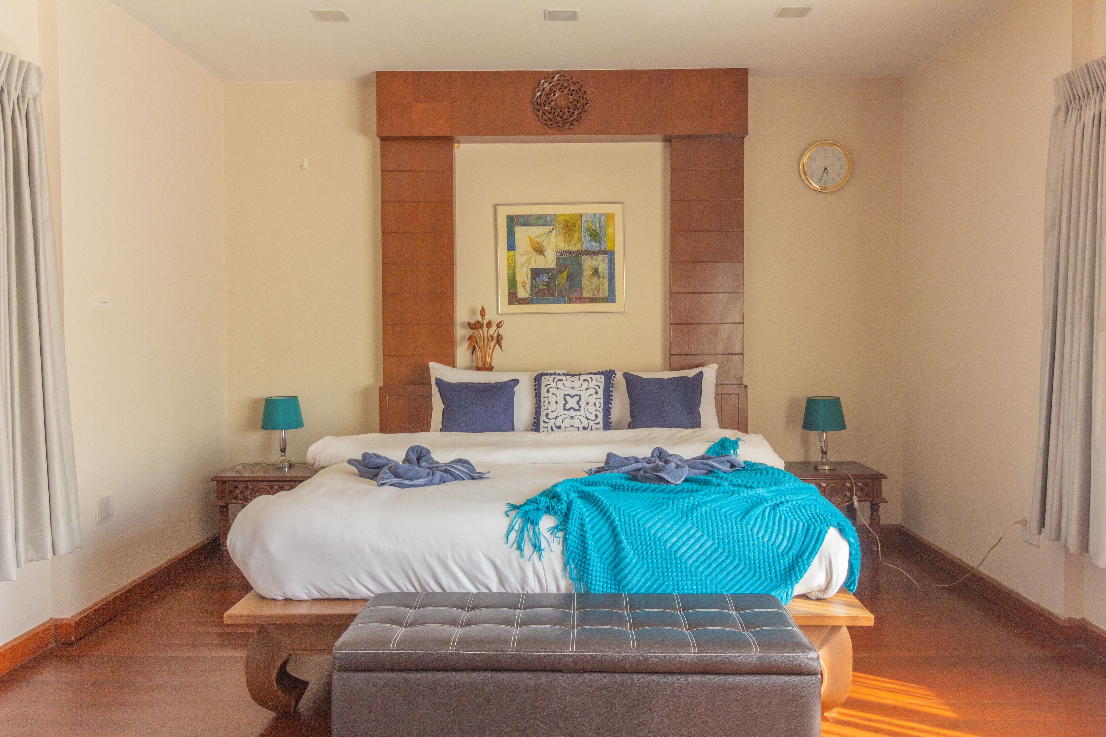 A bedroom with a large bed, two bedside tables with lamps, a wall clock, and a decorative piece above the bed.