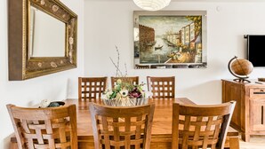A dining area with a wooden table, chairs, a vase of flowers, a painting of a canal scene, a mirror, and a globe on a cabinet. - Apartment 'Calonge Beach' with Private Terrace, Balcony and Air Conditioning (St Antoni de Calonge)