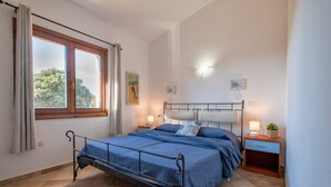 A bedroom with a metal bed frame, a nightstand with a lamp, a window with curtains, and a wall-mounted light fixture. - Holiday Home "Nuages" with Shared Pool and Private Terrace (Badesi)