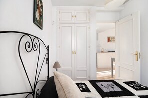 A bedroom with a bed, a white wardrobe, a door leading to a kitchen, and a wall-mounted picture.