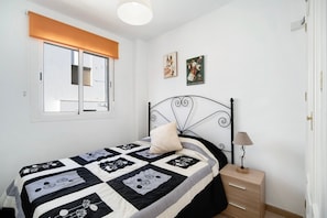 Une chambre à coucher comprenant un lit, une table de chevet, une lampe, une fenêtre avec des stores et deux œuvres d’art murales.