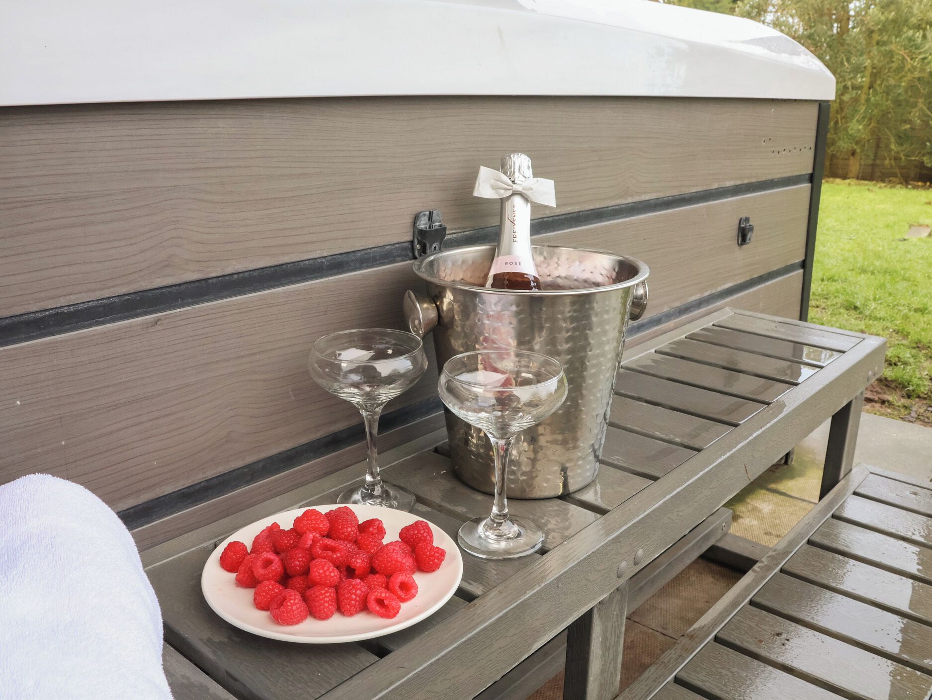 A picnic setup with a plate of raspberries, two wine glasses, and a champagne bottle in a metal bucket on a wooden table.