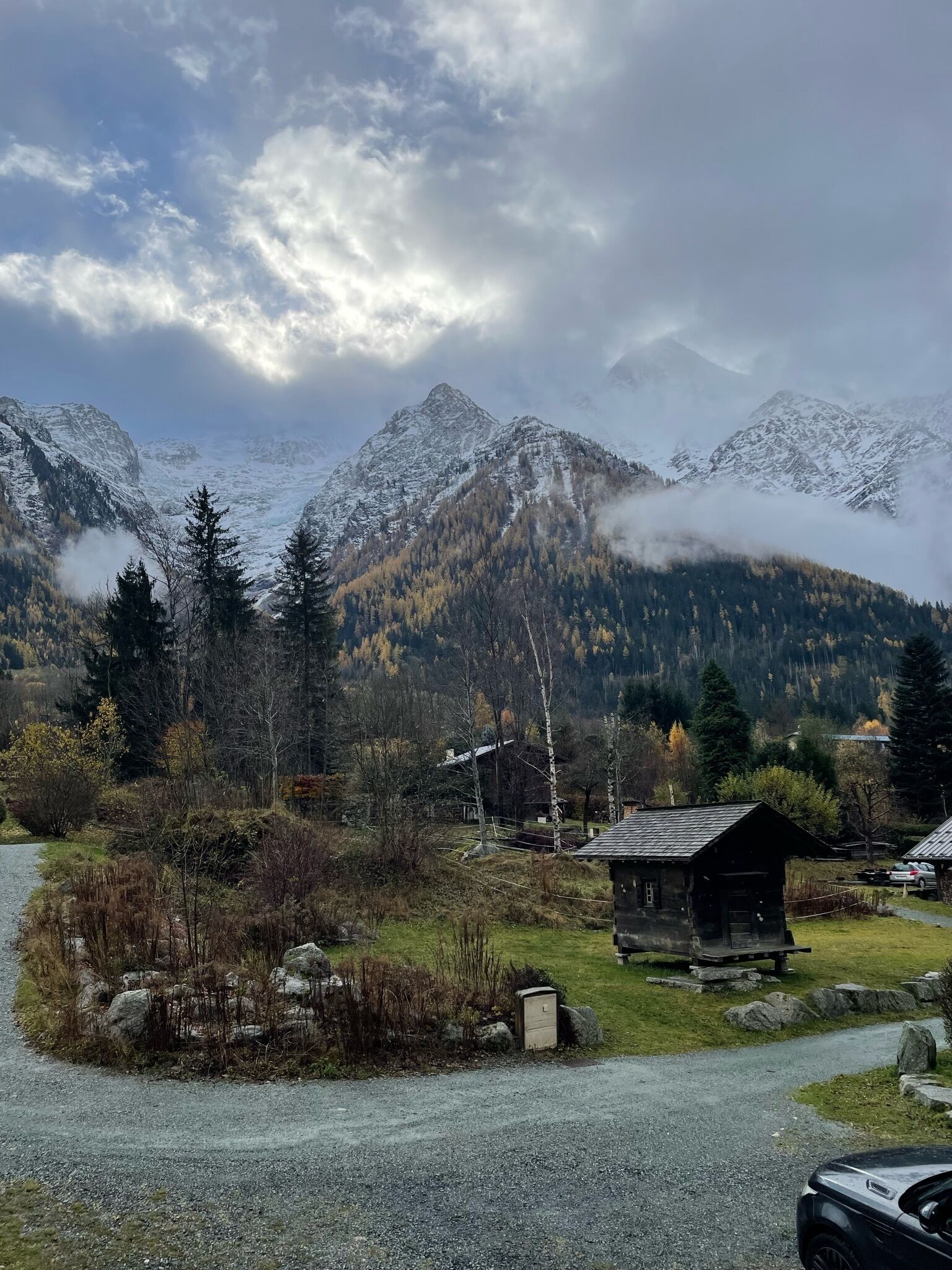 A rural mountain landscape with a small wooden cabin, a gravel path, and snow-capped mountains in the background.