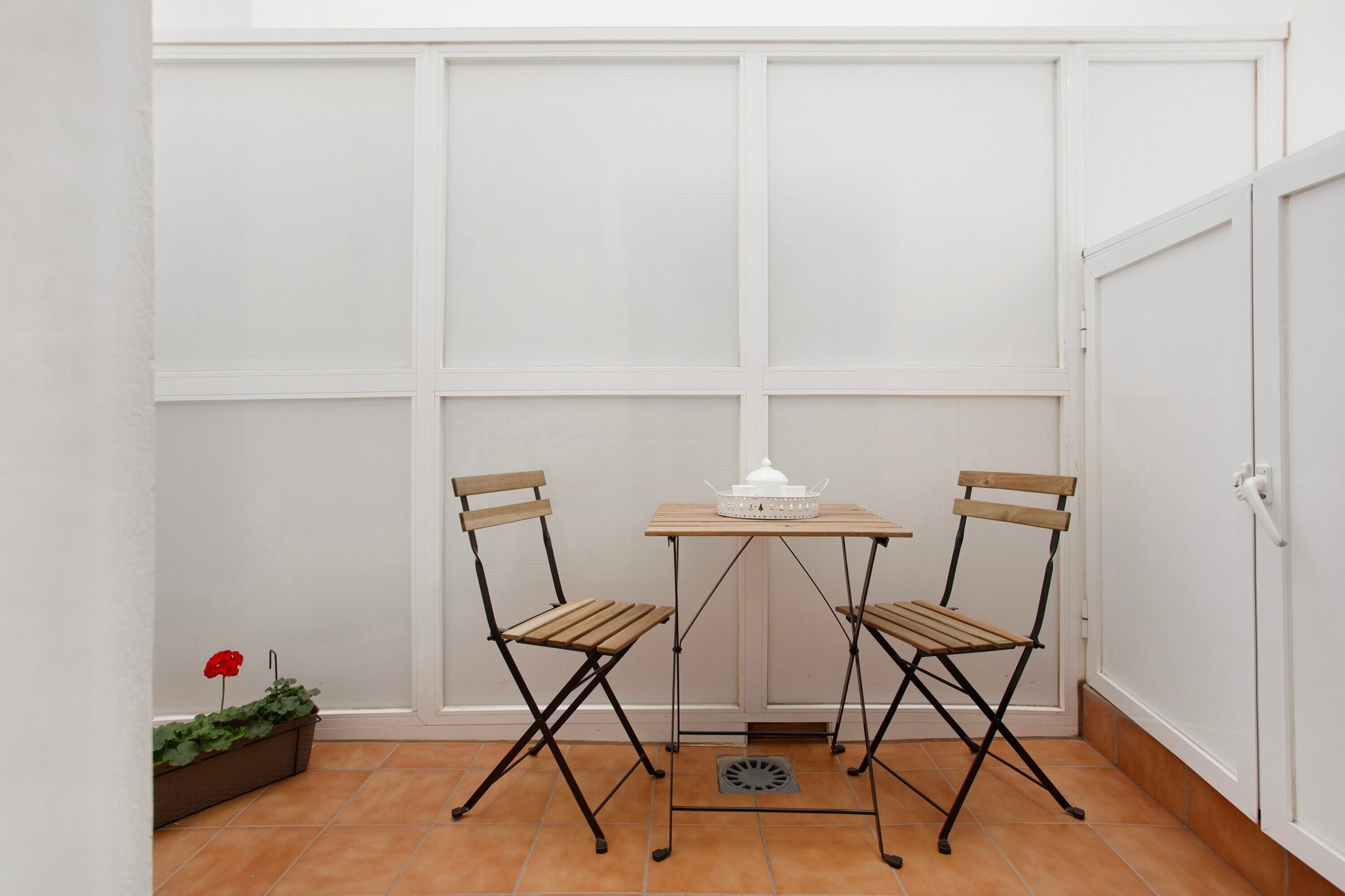 Una habitación pequeña con una puerta blanca, una ventana con contraventanas blancas, dos sillas de madera, una mesita con un objeto blanco y una planta en maceta con una flor roja.