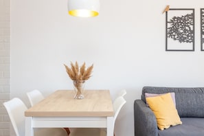 Une table à manger avec des chaises, un vase contenant des plantes séchées, un canapé gris avec un coussin jaune et un luminaire fixé au mur.