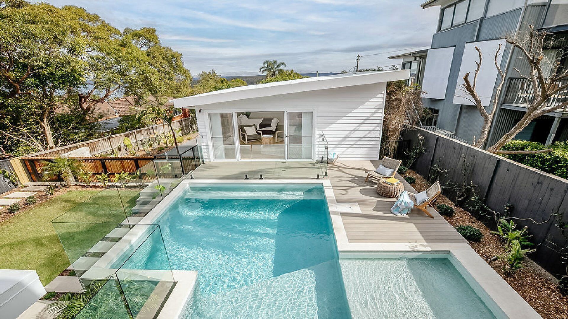 A modern backyard with a rectangular swimming pool, a covered patio area with seating, and a wooden deck.
