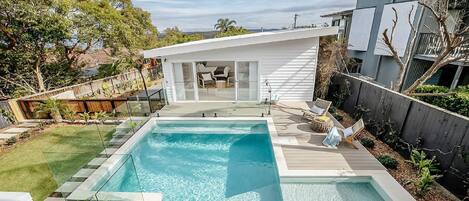 A modern backyard with a rectangular swimming pool, a covered patio area with seating, and a wooden deck.