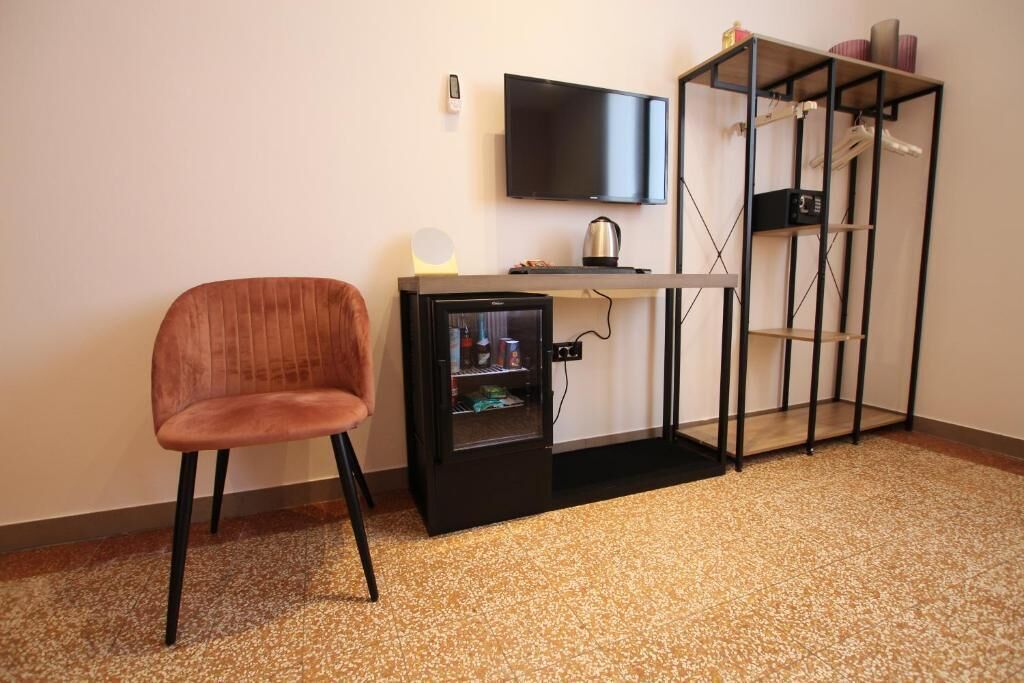 Um quarto com uma cadeira marrom, um pequeno armário preto com uma televisão montada acima e uma estante de metal com diversos objetos em cima.