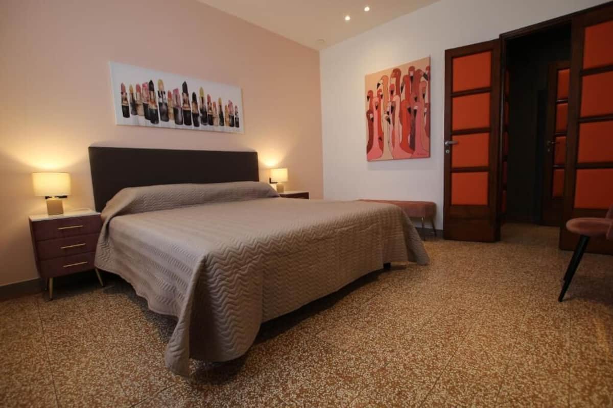Ein Schlafzimmer mit einem großen Bett, zwei Nachttischen mit Lampen, einer Tür mit rotem Rahmen und einer Wand mit zwei Kunstwerken.