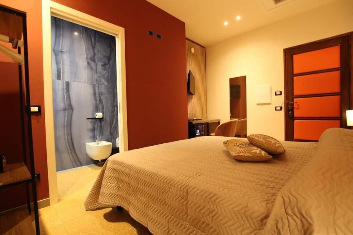 Ein Hotelzimmer mit Bett, Nachttisch, Stuhl, Spiegel, Fernseher und einem Badezimmer mit Waschbecken und Toilette.