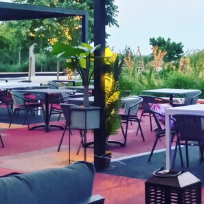 An outdoor seating area with tables and chairs, surrounded by greenery and decorative lights.