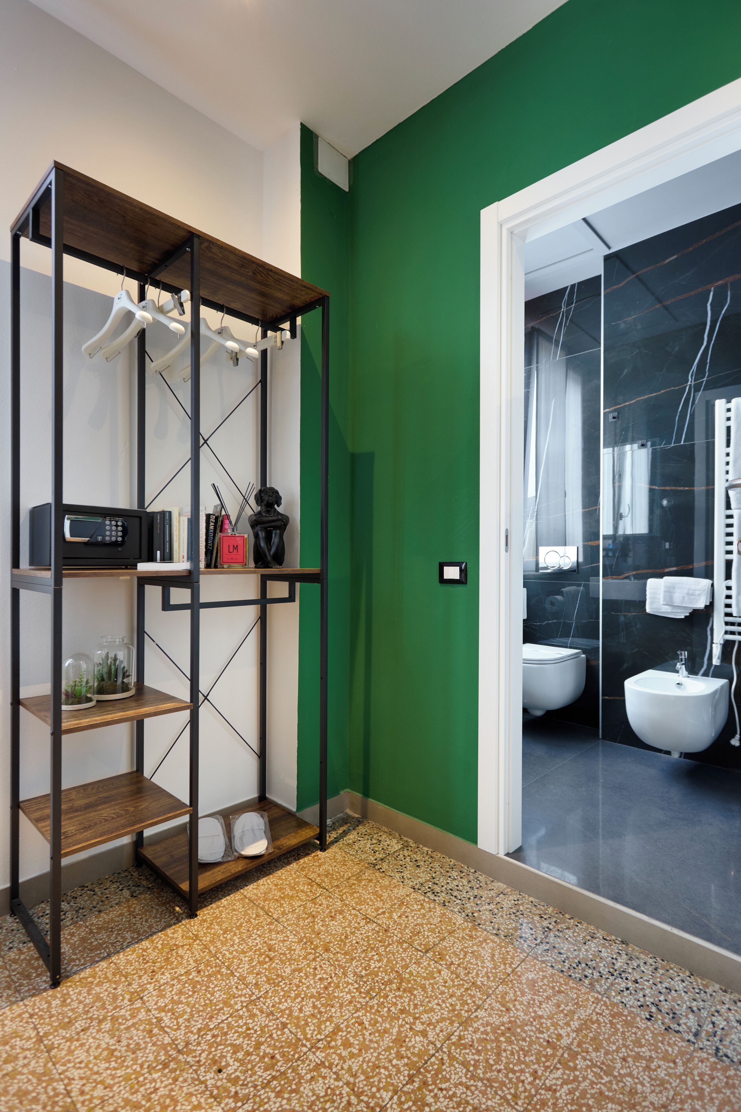 Una habitación con pared verde, una estantería de madera con objetos colgantes, una puerta blanca que da a un baño y suelo de baldosas.