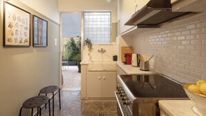 A compact kitchen with a stove, sink, and a window with blinds. There are two stools and framed pictures on the wall. - Marina Zeas Apartment by the sea (Piraeus)