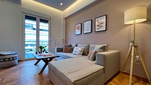 A modern living room with a sofa, a coffee table, and a floor lamp. There are framed pictures on the wall and a window with a view of the outside. - Nea Smirni Square Chic Apartment (Nea Smirni)