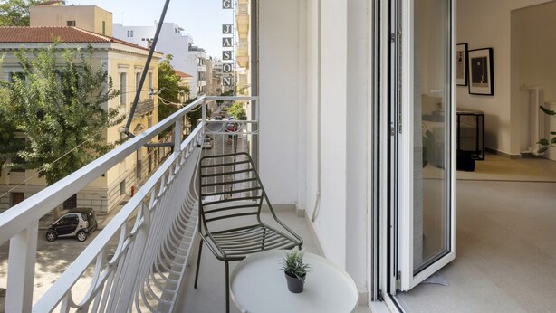 En balkon med en metalstol og et lille bord, en potteplante samt udsigt til gaden og bygningerne.