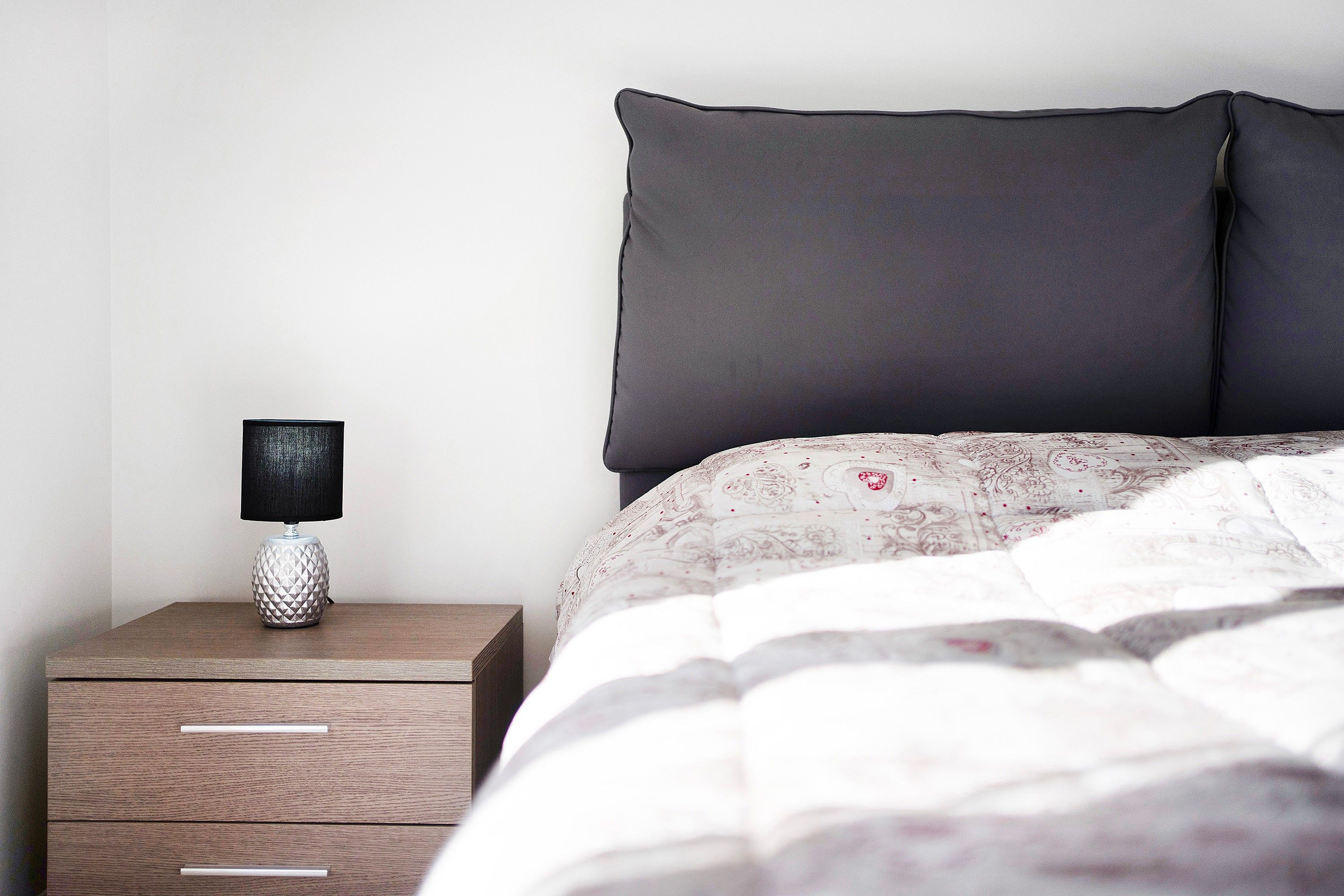Een netjes opgemaakt bed met een bedsprei met dessin, een bedlampje op een houten nachtkastje en een effen muur op de achtergrond.