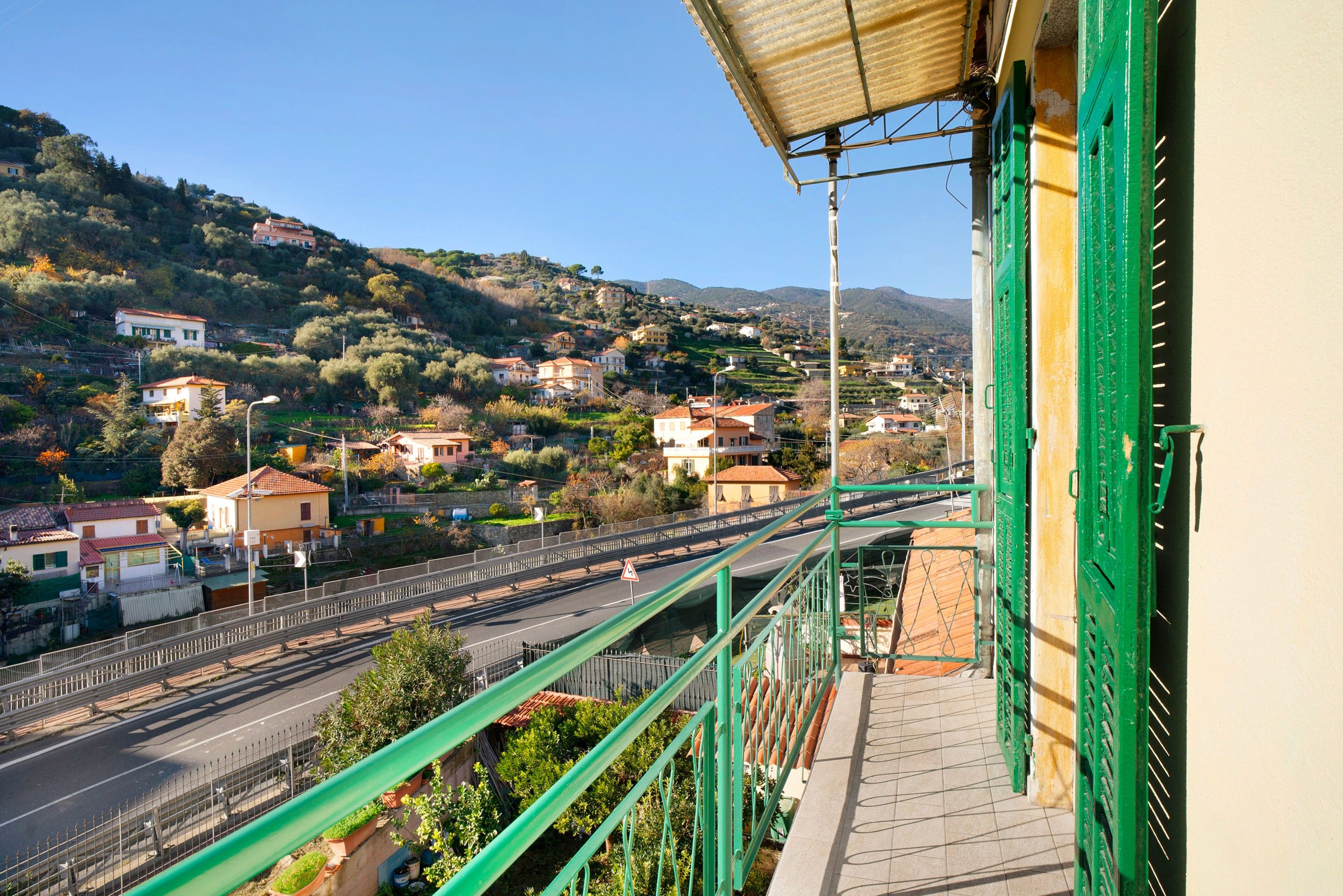 Un balcon donnant sur une ville située sur une colline, une route avec un pont piétonnier et une rambarde métallique verte.