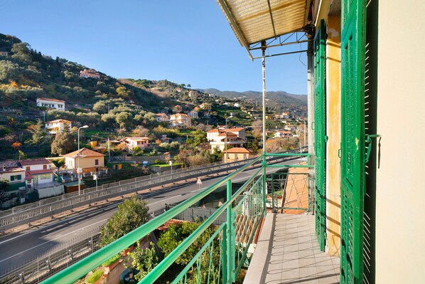 A balcony with a view of a hillside town, a road with a pedestrian bridge, and a green metal railing.