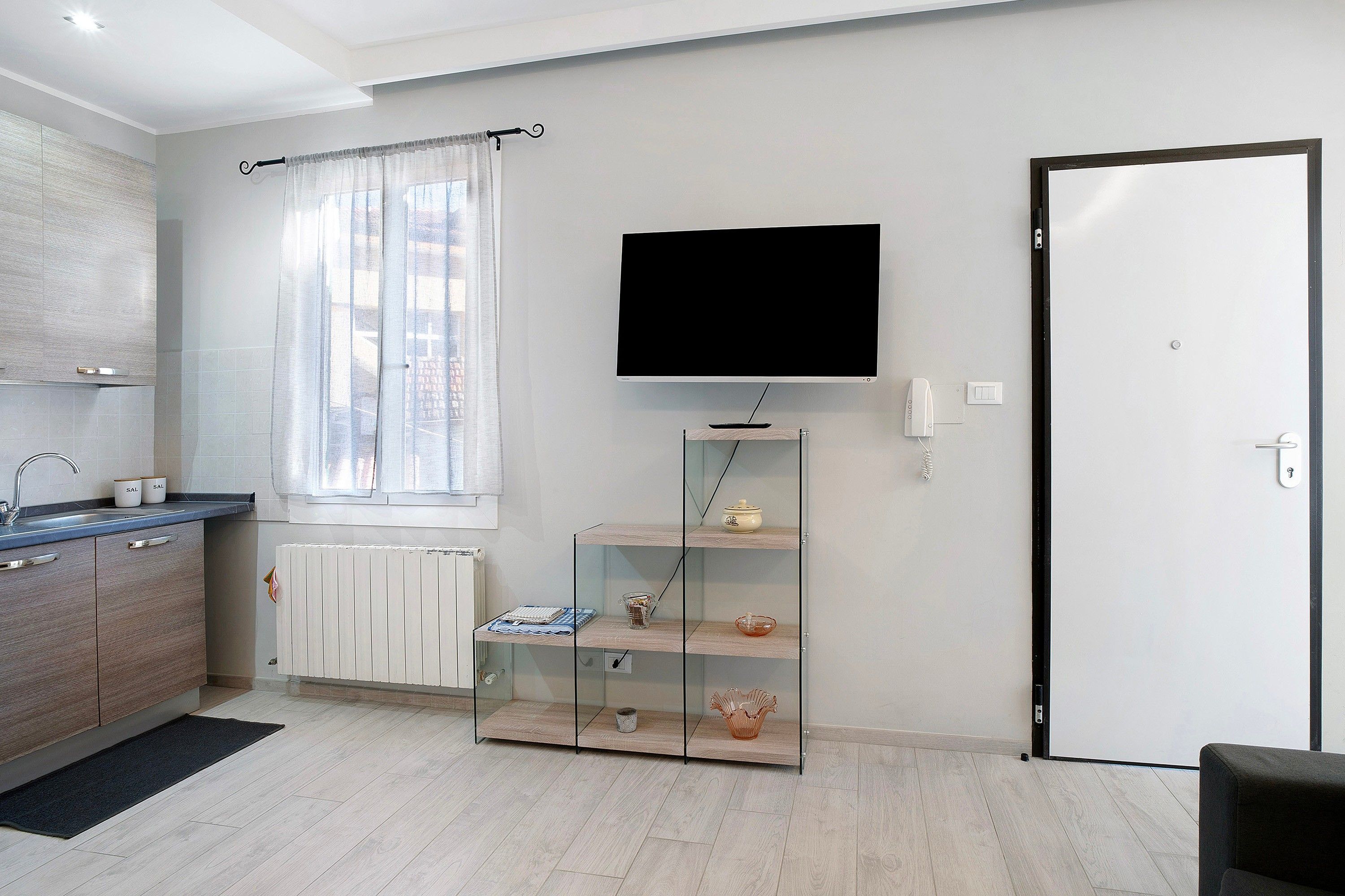 A modern kitchen with a built-in oven, a window with curtains, a flat-screen TV mounted on the wall, and a door with a handle.