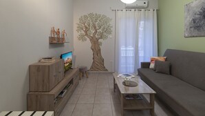 A living room with a wooden TV stand, a coffee table, a grey sofa, a wall-mounted air conditioner, and a large tree mural. - Cozy Athenian 1br: Pistachio Charm (Athens)