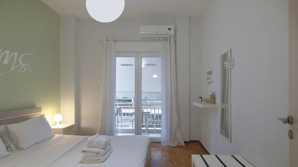 A hotel room with a bed, a bedside table, a window with curtains, a view of the outside balcony, and an air conditioning unit. - Cozy Athenian 1br: Pistachio Charm (Athens)