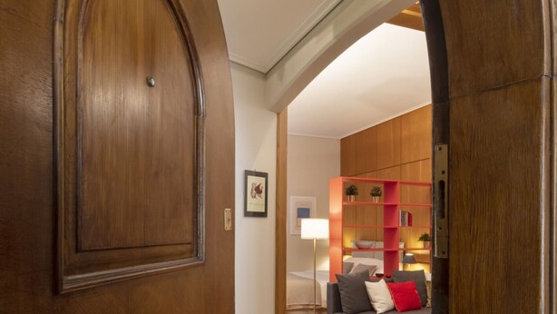 Uma porta de madeira dá acesso a uma sala de estar com um sofá, uma estante vermelha e um abajur.