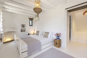 A bedroom with a white stone wall, a bed with a wooden headboard, a nightstand, a small wooden cabinet, and a hanging light fixture.