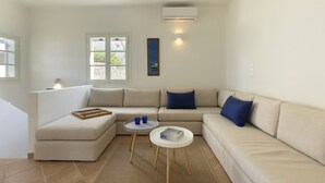 A modern living room with a beige sectional sofa, two small round tables, and a large window with a view of greenery. - Stunning Beachfront 3BR Villa (Paros)