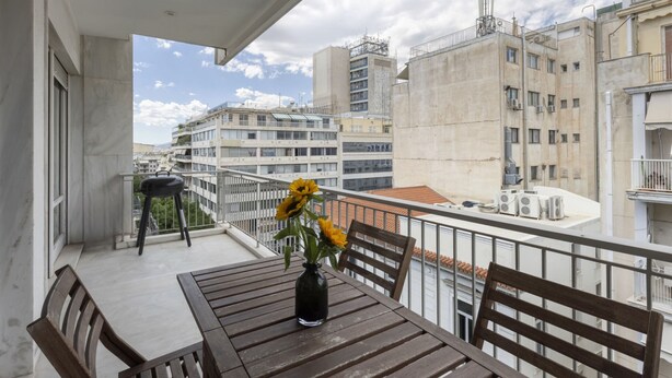 Ein Balkon mit Holztisch und -stühlen, einer Vase mit gelben Blumen und Blick auf umliegende Gebäude.