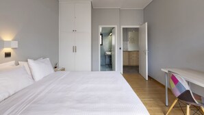 Ein Schlafzimmer mit einem großen Bett, einem Nachttisch, einem Schreibtisch und einem Stuhl. Ein Kleiderschrank ist zu sehen, und durch eine offene Tür gelangt man ins Badezimmer.